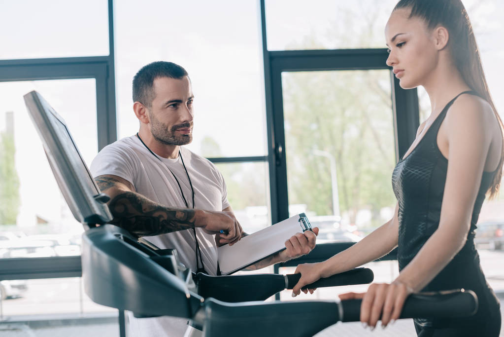 male personal trainer holding clipboard and sportswoman looking at treadmill screen  - Photo, Image