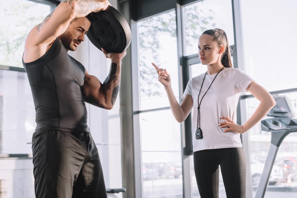 entrenadora personal femenina contando mientras deportista haciendo ejercicios con disco de barra en el gimnasio
 - Foto, imagen