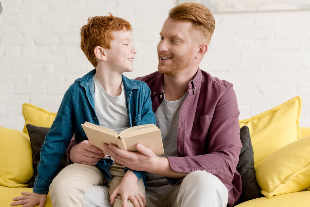 onnellinen isä ja poika hymyilevät toisiaan lukiessaan kirjaa yhdessä kotona
 - Valokuva, kuva