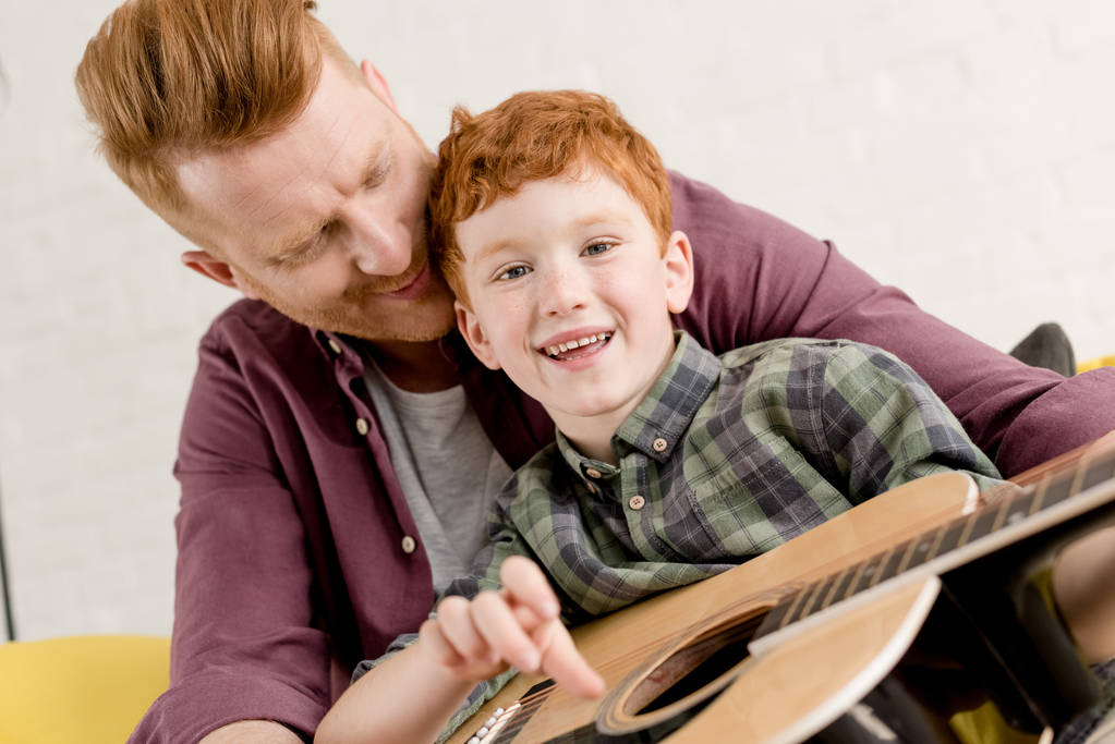 ευτυχισμένος/η κοκκινομάλλης/α πατέρας και γιος που παίζουν ακουστική κιθάρα μαζί στο σπίτι - Φωτογραφία, εικόνα