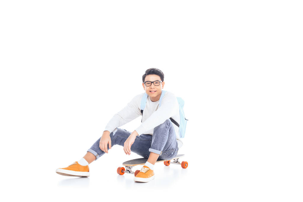 asiático adolescente con mochila sentado en skateboard aislado en blanco
 - Foto, imagen