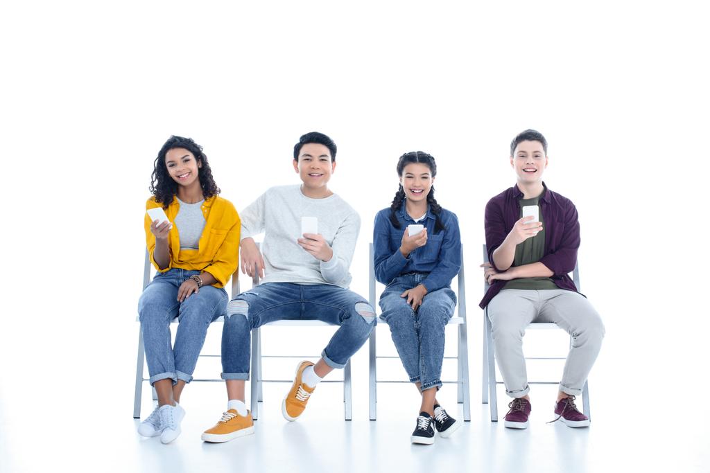 multi-etnisch tiener studenten met smartphones zitten op stoelen geïsoleerd op wit - Foto, afbeelding