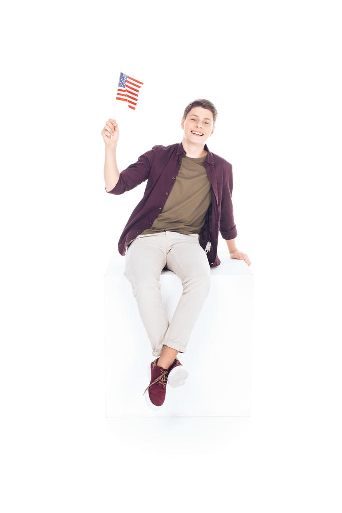 adolescente estudante menino sentado no cubo branco e olhando para a câmera com bandeira EUA isolado no branco
 - Foto, Imagem