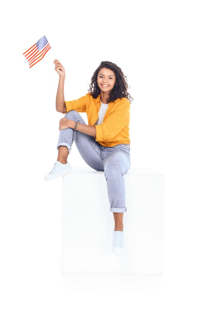nastoletnich african american student dziewczyna siedzi na białego sześcianu i patrząc na kamery z usa bandery na białym tle - Zdjęcie, obraz