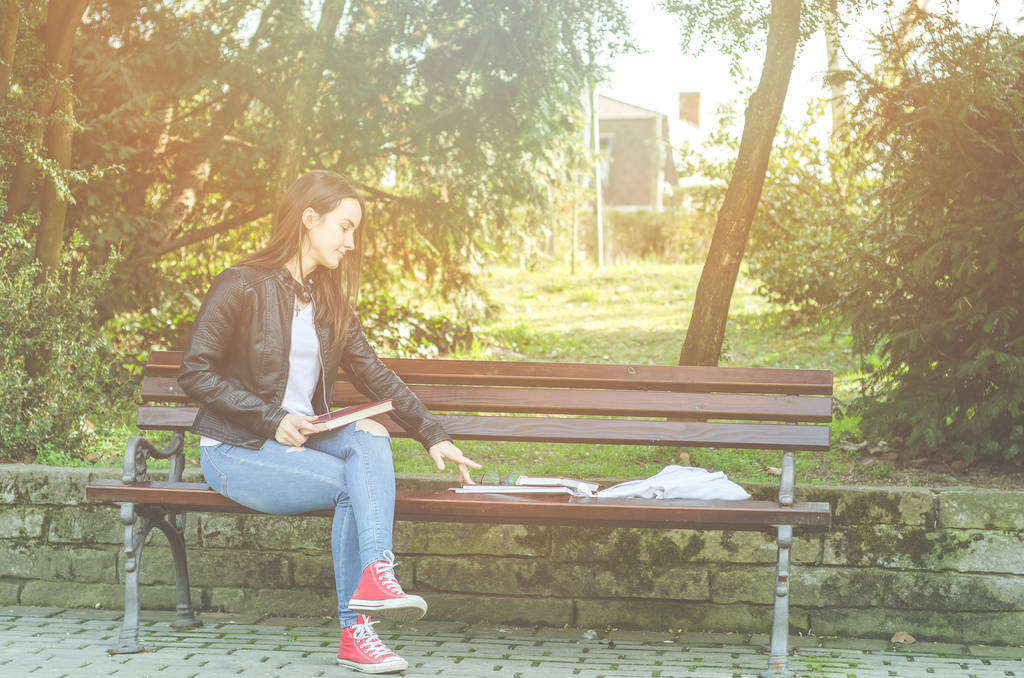 Young beautiful school or college girl with eyeglasses sitting on the bench in the park reading the books and study for exam, education concept - Photo, Image