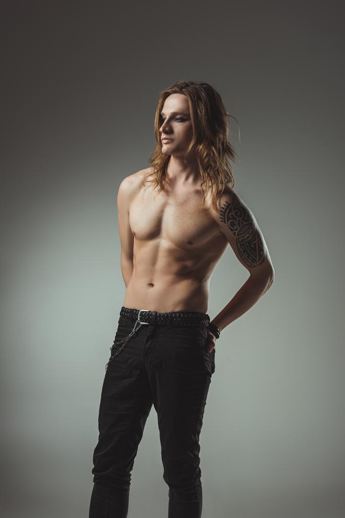 Мужчина без рубашки с длинными волосами и татуировкой, позирующий в черных джинсах, изолированный на сером
 - Фото, изображение