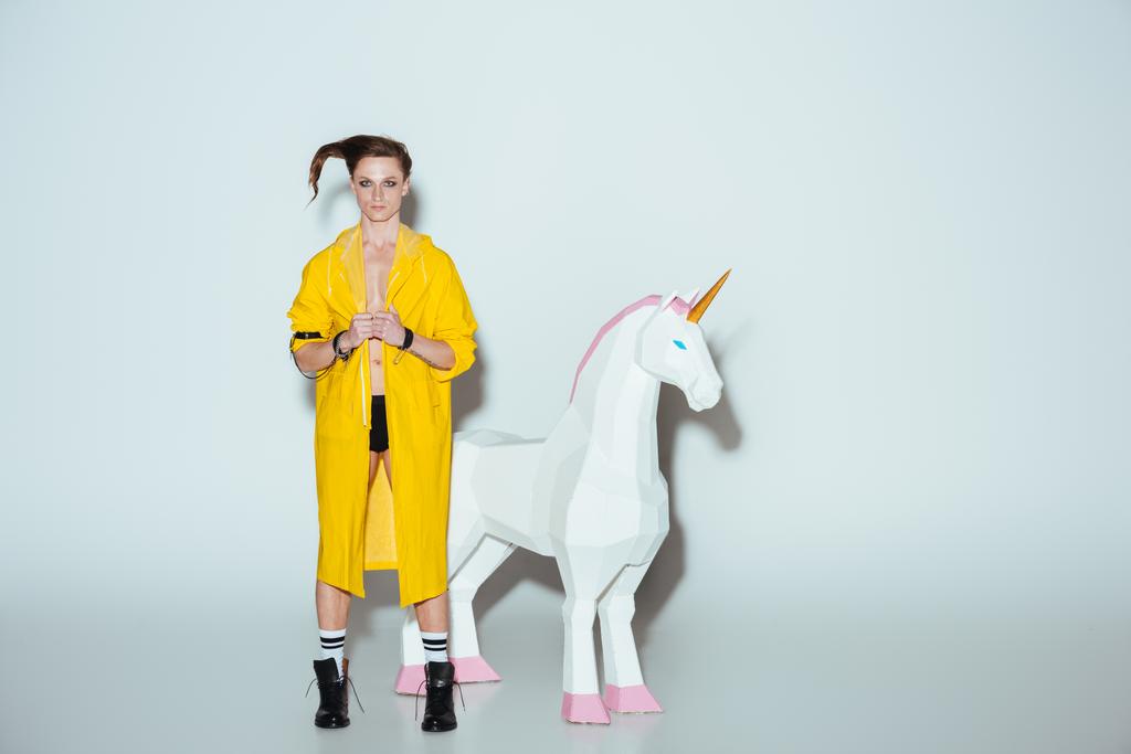 fashionable man with hairstyle in yellow raincoat standing with big unicorn toy, on grey - Photo, Image