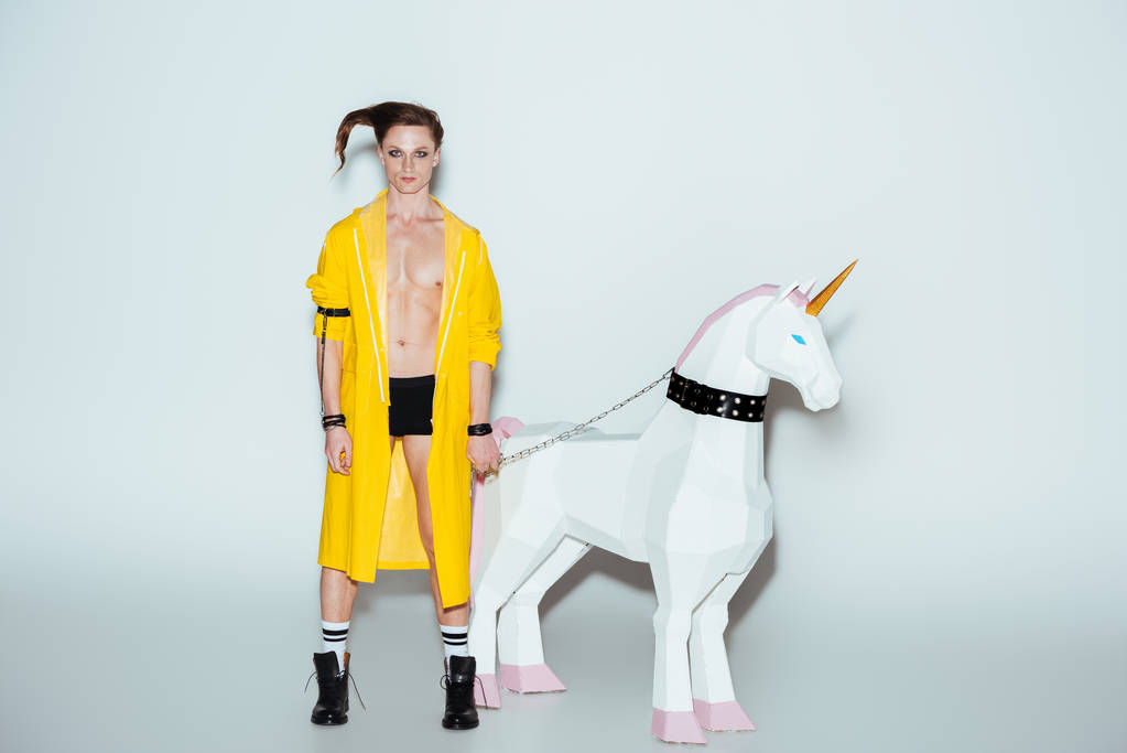 mannelijk model in boxershorts en gele regenjas permanent met grote unicorn speelgoed op de ketting, grijs - Foto, afbeelding