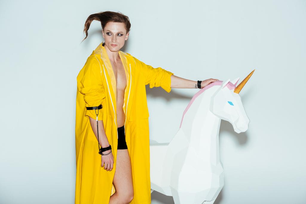 man with strange hairstyle in yellow raincoat standing with big unicorn toy, on grey - Photo, Image