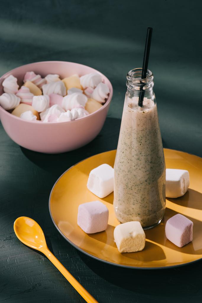 Nahaufnahme von Milchshake in Flasche mit Trinkhalm, Schüssel mit Marshmallows und Löffel auf Tischplatte - Foto, Bild