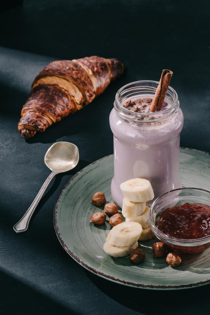 Close-up shot van berry smoothie met kaneel stro en chocolade schaafsel, walnoten, jam en banaan segmenten op de plaat, lepel en croissant  - Foto, afbeelding