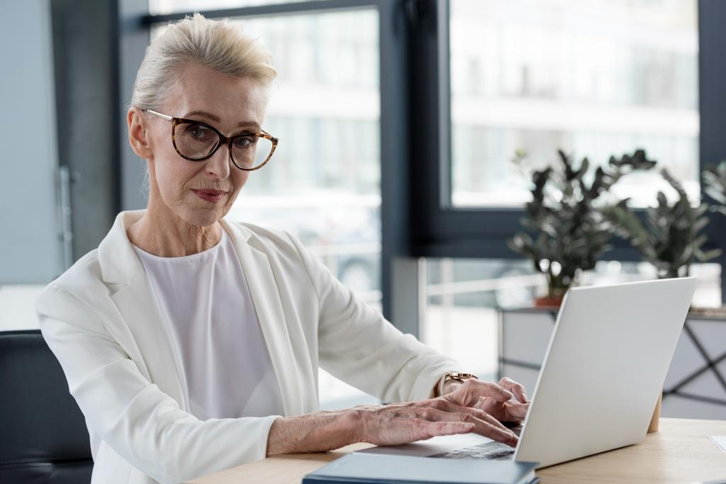 hermosa mujer de negocios senior en gafas mirando a la cámara mientras se utiliza el ordenador portátil en el lugar de trabajo
 - Foto, Imagen