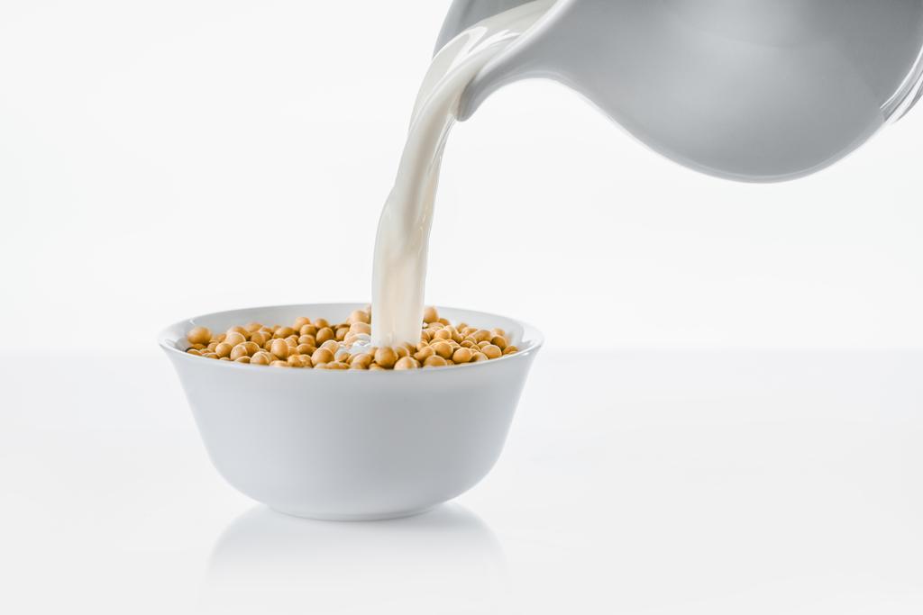 Milch aus Krug in Schüssel mit Sojabohnen auf weißem Hintergrund gießen - Foto, Bild