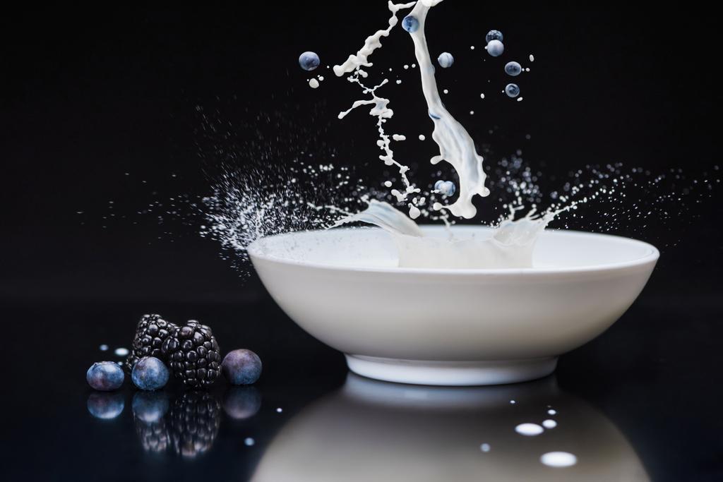 Fresh berries splashing in white bowl with milk on black background - Photo, Image