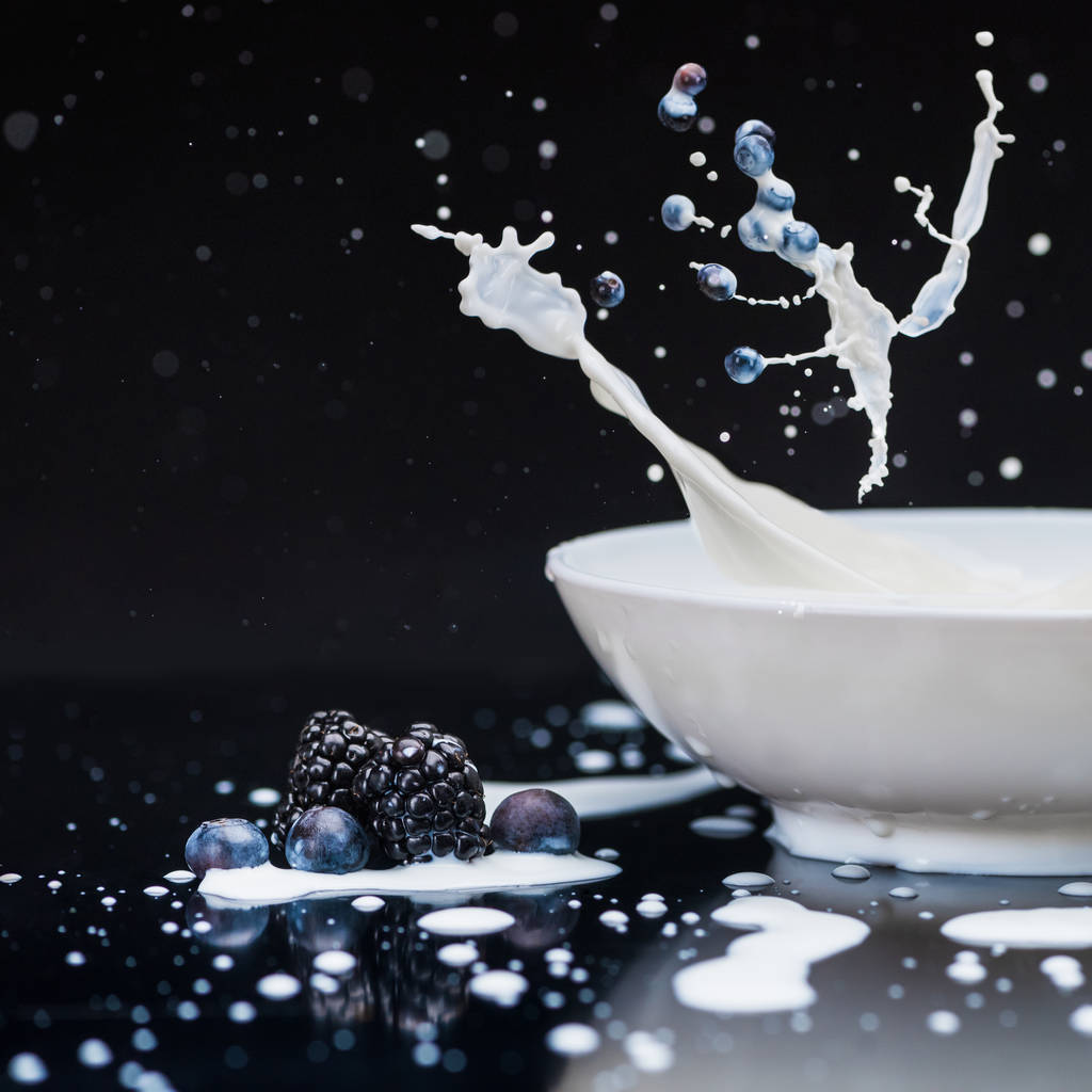 Sappige bessen vallen in witte kom met spatten van melk op zwarte achtergrond - Foto, afbeelding