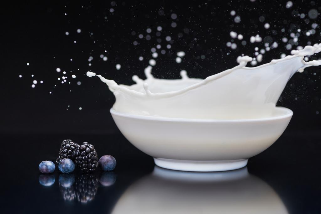 saftige Beeren von weißen Schüssel mit Spritzmilch auf schwarzem Hintergrund - Foto, Bild