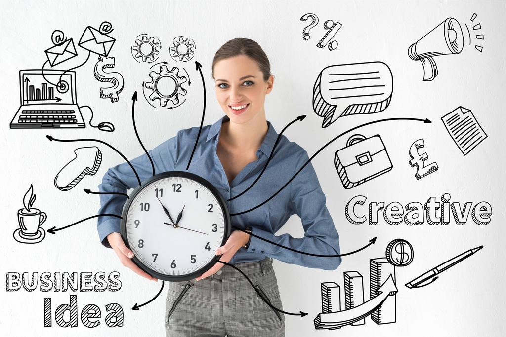 Usměvavá podnikatelka drží nástěnné hodiny v rukou na bílém s kreativní firmy idea nápis a ručně kreslené ikony - Fotografie, Obrázek