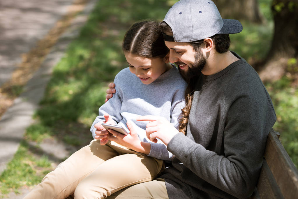 pai feliz e filha usando smartphone no banco no parque
 - Foto, Imagem