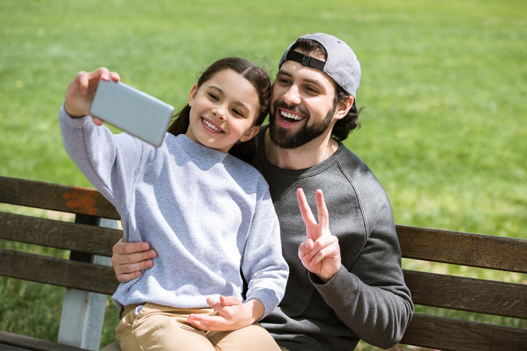 dochter nemen selfie met vader doet vrede gebaar in park  - Foto, afbeelding
