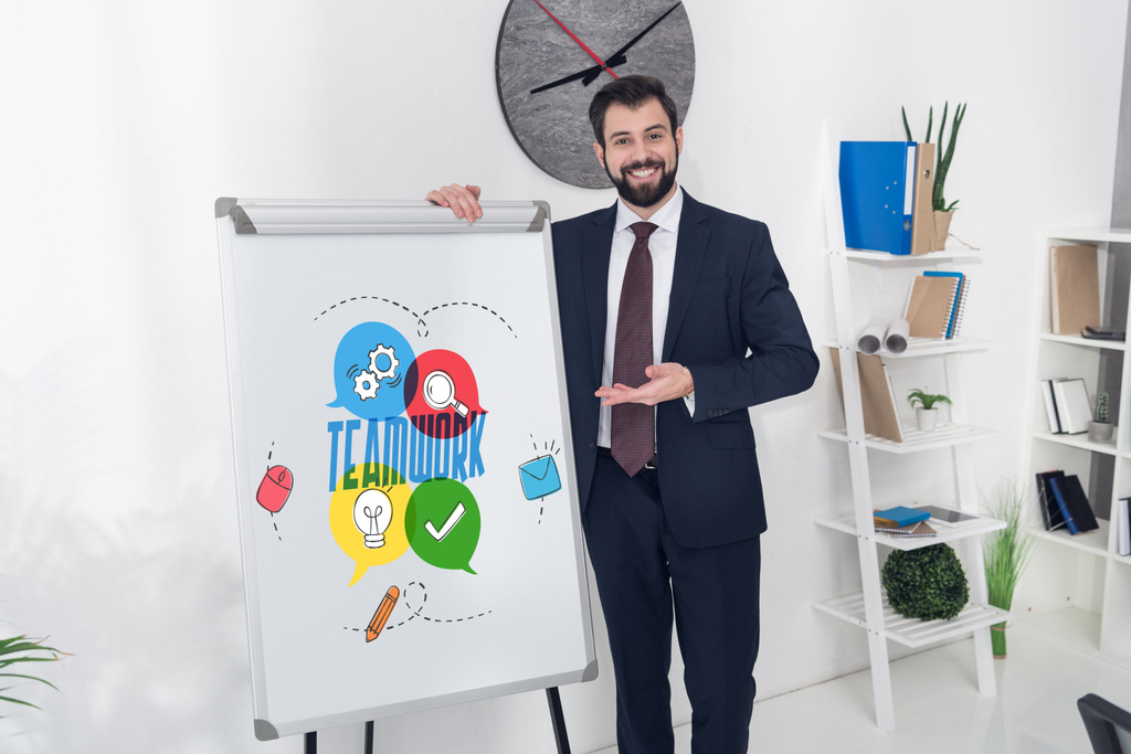 Улыбающийся бизнесмен, указывающий на белую доску с надписью "командная работа" и деловыми иконами в офисе
 - Фото, изображение