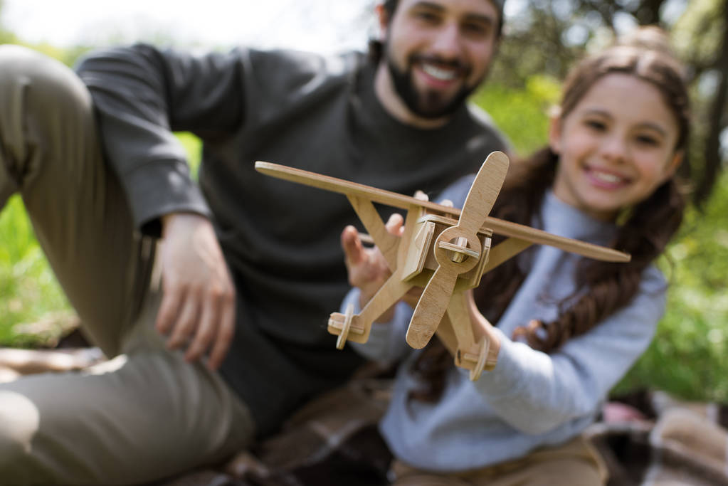 vista ravvicinata di aereo giocattolo di legno in mani di ragazza seduta su plaid con padre in parco
  - Foto, immagini