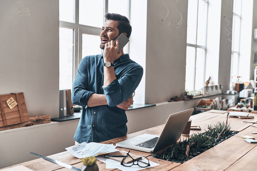 glücklicher schöner junger Mann, der am Holztisch mit Laptop im modernen Büro steht und mit dem Smartphone telefoniert - Foto, Bild
