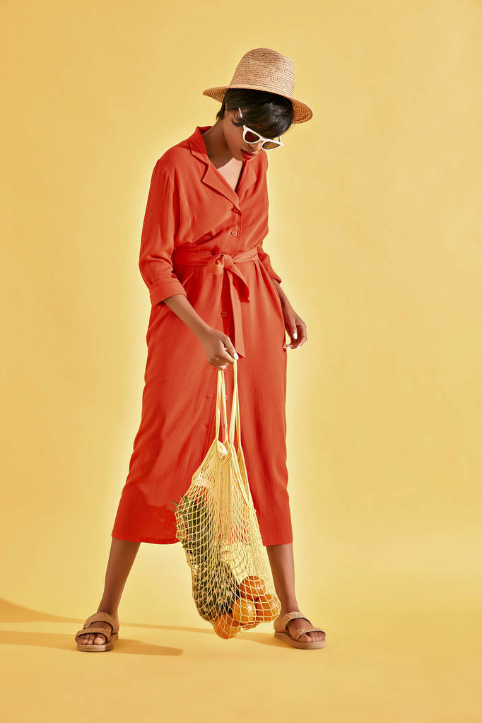 Barevný portrét mladá Černoška s hnědými vlasy a červené rty, oblečený v oranžové šaty a slaměný klobouk a boty s ovocem do čistého sáčku. Studio záběr, žluté pozadí - Fotografie, Obrázek