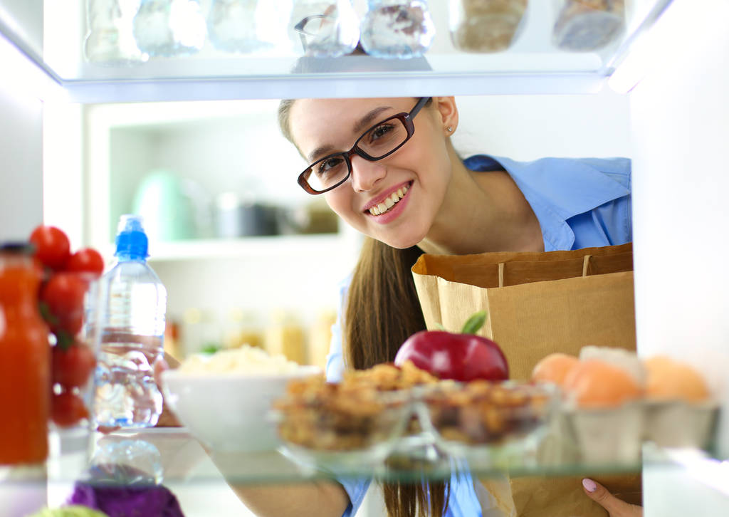Portret van een vrouw in de buurt van een open koelkast vol gezond voedsel, groenten en fruit - Foto, afbeelding