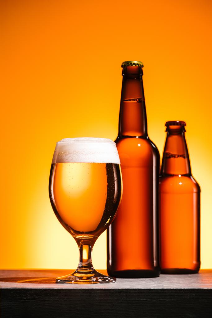 vista de perto de garrafas e vidro de cerveja com espuma na superfície sobre fundo laranja
 - Foto, Imagem