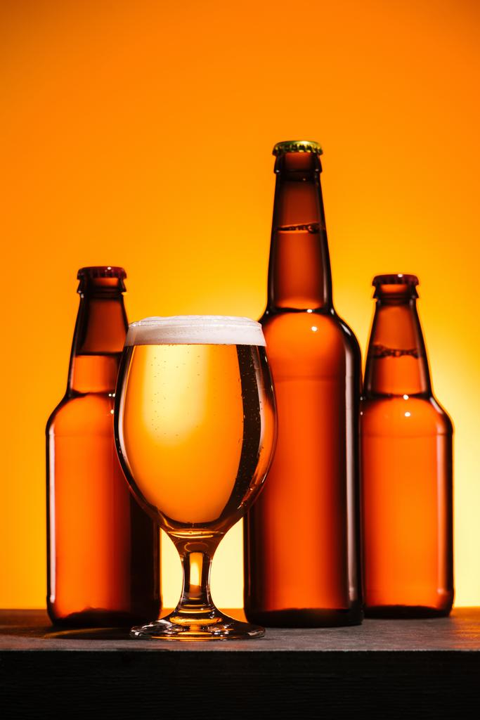 κλείνω πάνω θέα φιαλών και ποτήρι μπύρα με αφρό στην επιφάνεια σε πορτοκαλί φόντο - Φωτογραφία, εικόνα