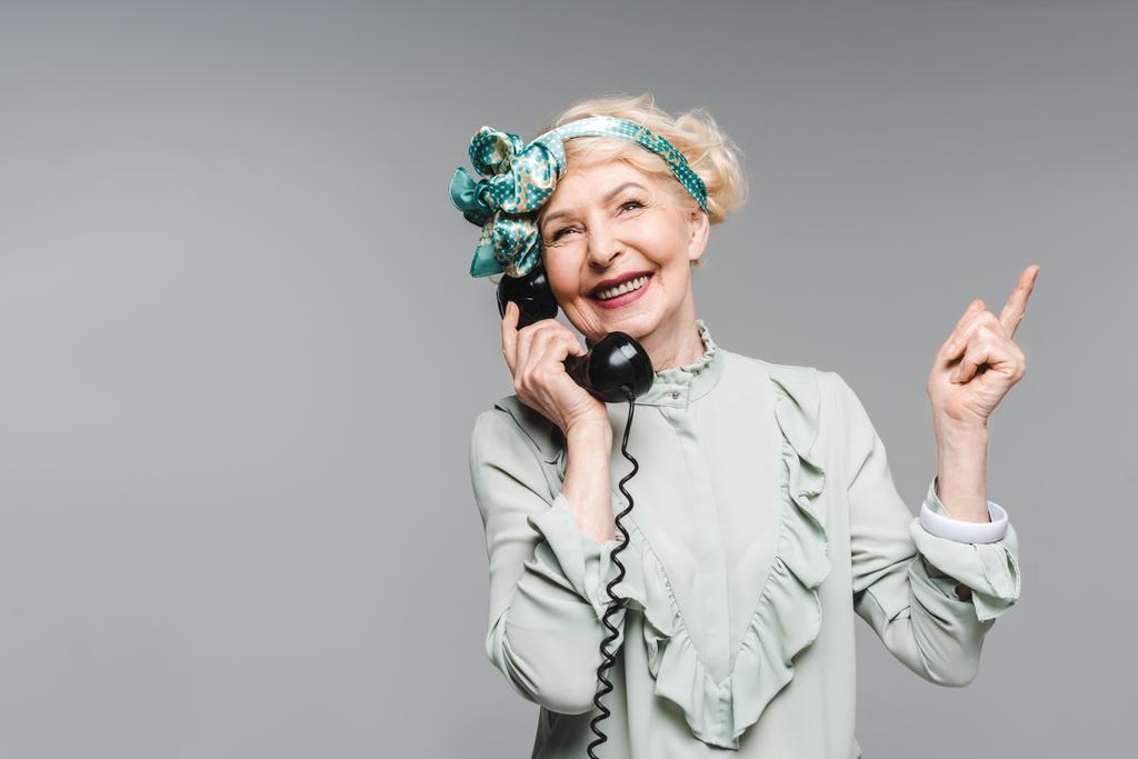 feliz mulher sênior falando por telefone vintage e apontando para cima isolado em cinza
 - Foto, Imagem