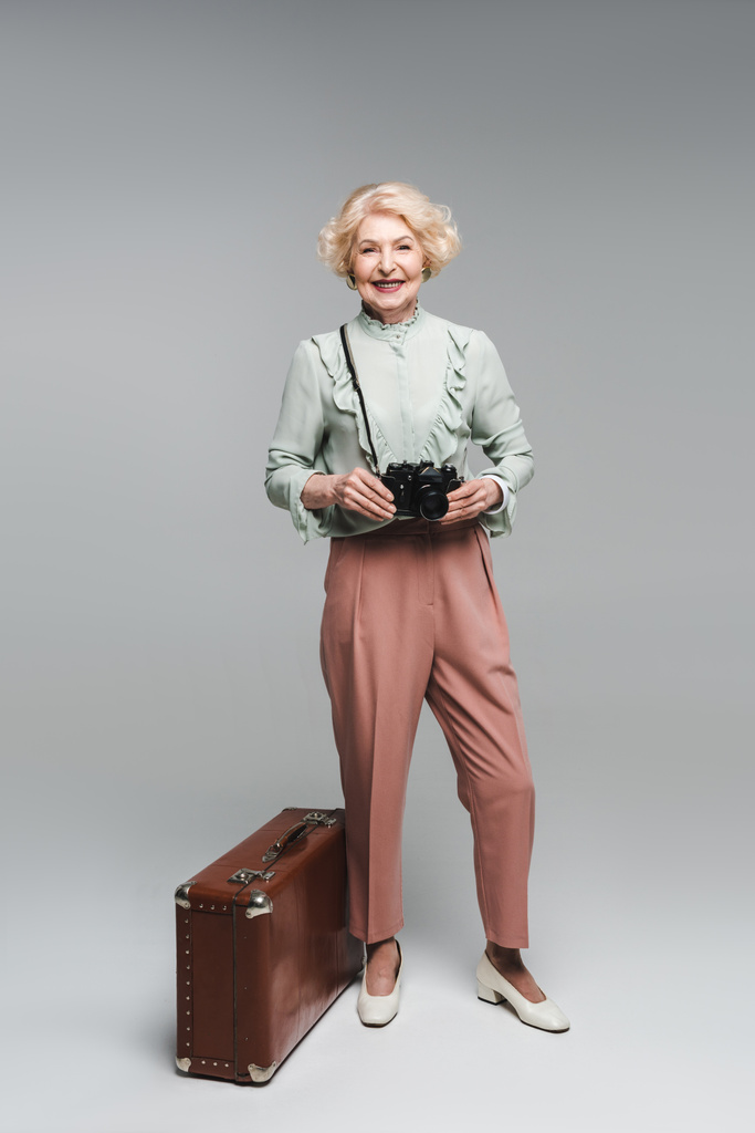 όμορφη γυναίκα ανώτερος με vintage βαλίτσα και φιλμ κάμερας σε γκρι - Φωτογραφία, εικόνα