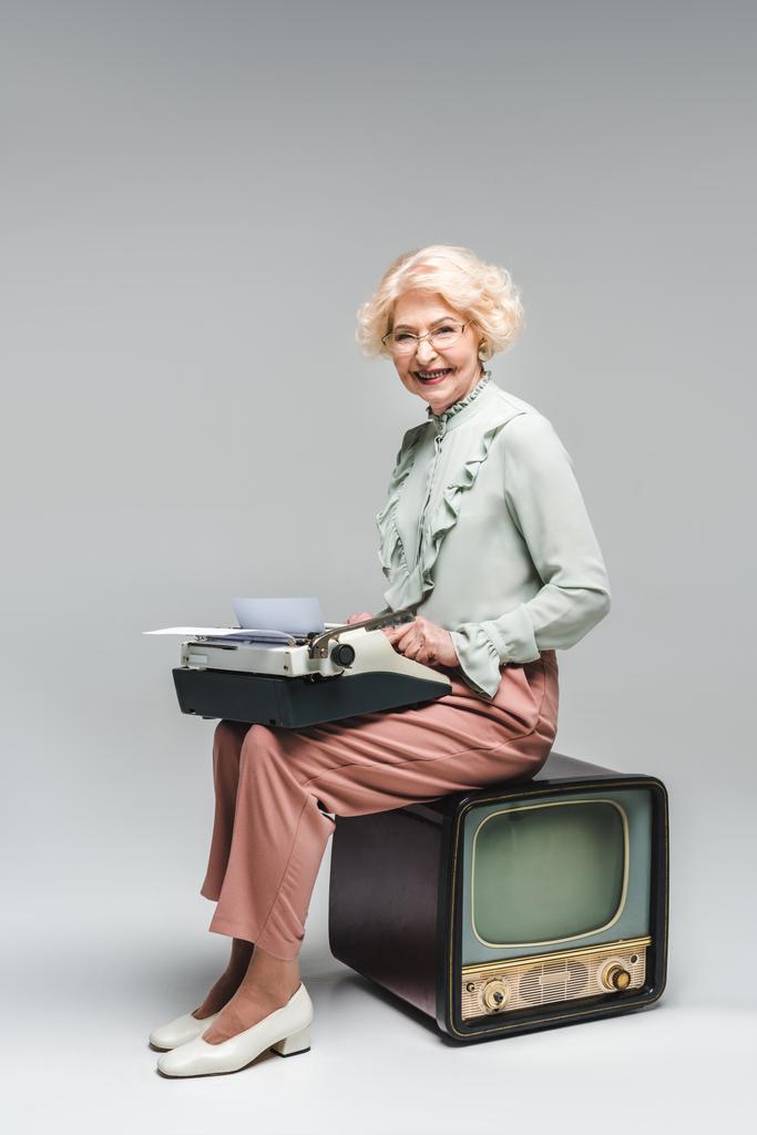 usměvavá starší žena pomocí psacího stroje, zatímco sedí na vintage tv Grey - Fotografie, Obrázek