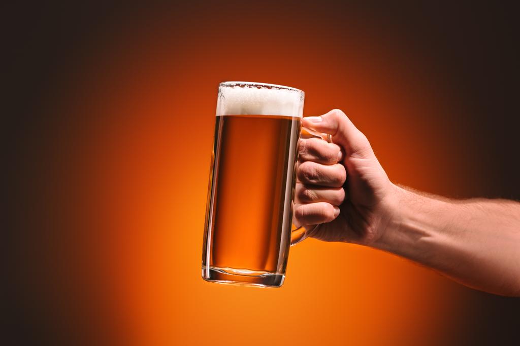 περικοπεί shot του άνδρα να κρατάει την κούπα της κρύας μπύρας σε πορτοκαλί φόντο - Φωτογραφία, εικόνα