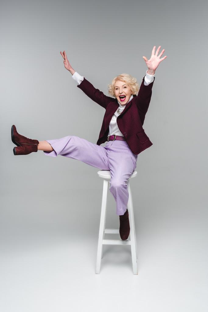 кричуща смішна старша жінка сидить на стільці з піднятими руками і ногами на сірому
 - Фото, зображення