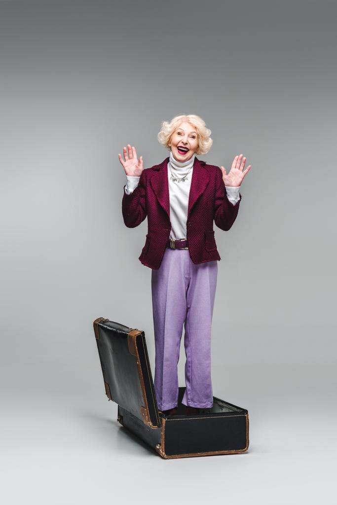 happy senior woman standing inside of vintage suitcase with raised hands on grey - Photo, Image