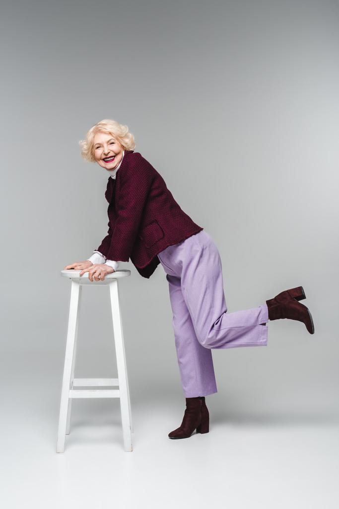 schöne Seniorin in stilvoller Vintage-Kleidung lehnt auf Stuhl auf grau - Foto, Bild