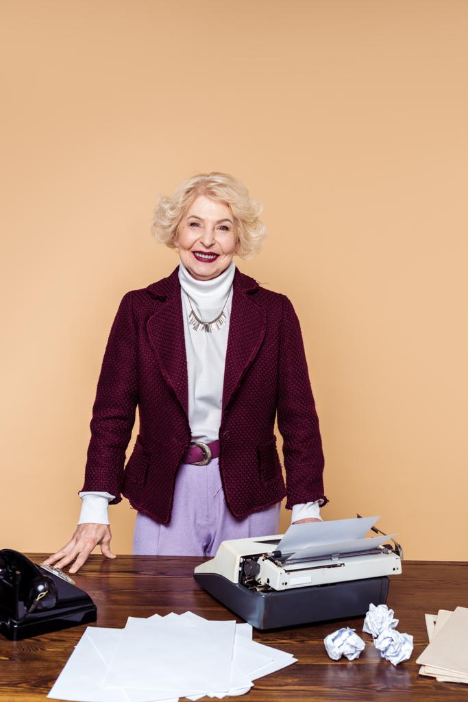 uśmiechający się stylowe starszy kobieta stojąc przy stole z telefonu do pisania i obrotowe - Zdjęcie, obraz