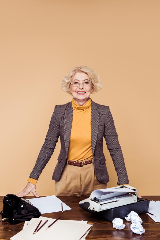 Lächelnde Seniorin mit Brille, die mit Papieren, Schreibmaschine und Telefon am Tisch steht - Foto, Bild