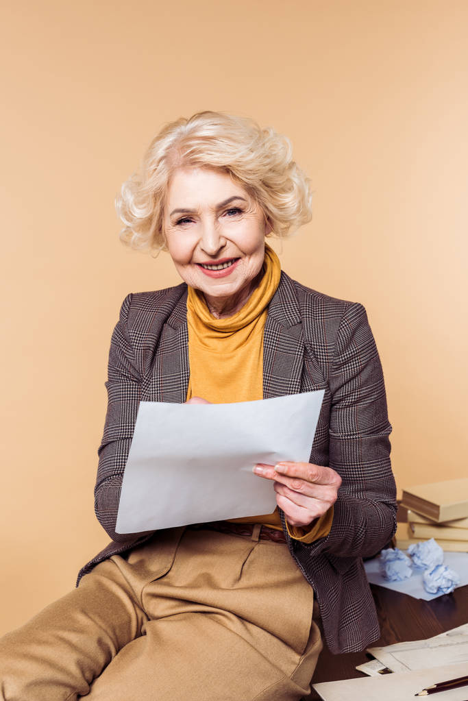 uśmiechający się starszy kobieta siedzi przy stole z papieru na białym tle na beżowym tle  - Zdjęcie, obraz