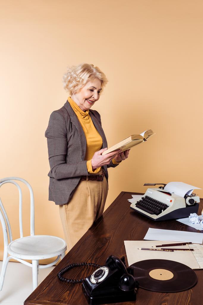 sorridente sênior mulher ler livro perto da mesa com telefone rotativo, disco de vinil e máquina de escrever
  - Foto, Imagem