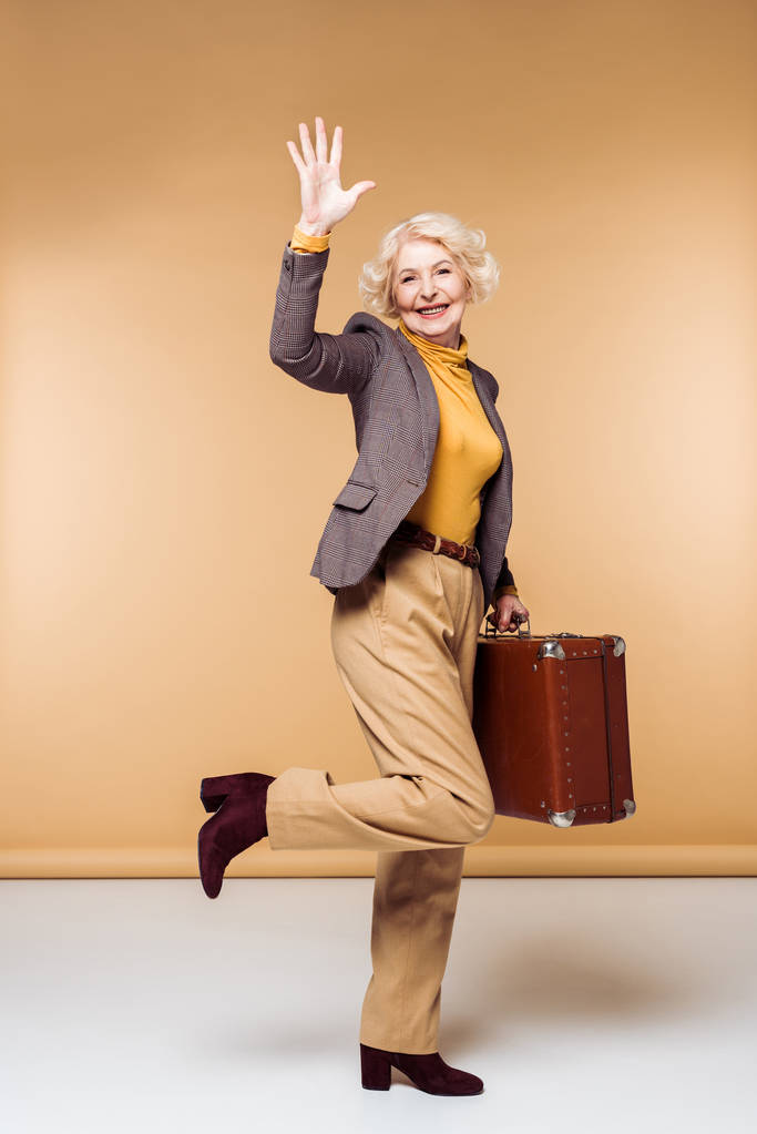 stijlvolle senior vrouwelijke reiziger zwaaien met de hand en houden van vintage koffer  - Foto, afbeelding