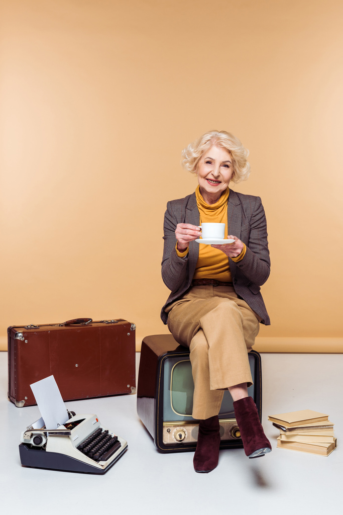 κομψή ανώτερων γυναίκα πίνοντας καφέ και κάθεται στο vintage tv κοντά σε γραφομηχανή και βαλίτσα  - Φωτογραφία, εικόνα