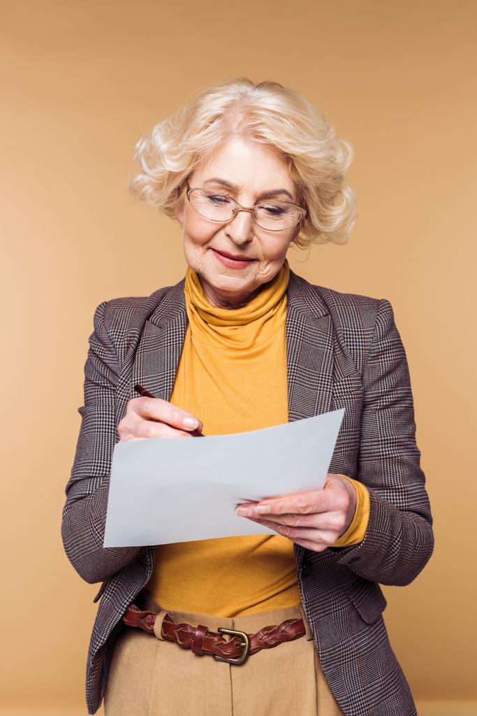stylische Seniorin mit Brille, die auf Papier auf beigem Hintergrund schreibt  - Foto, Bild