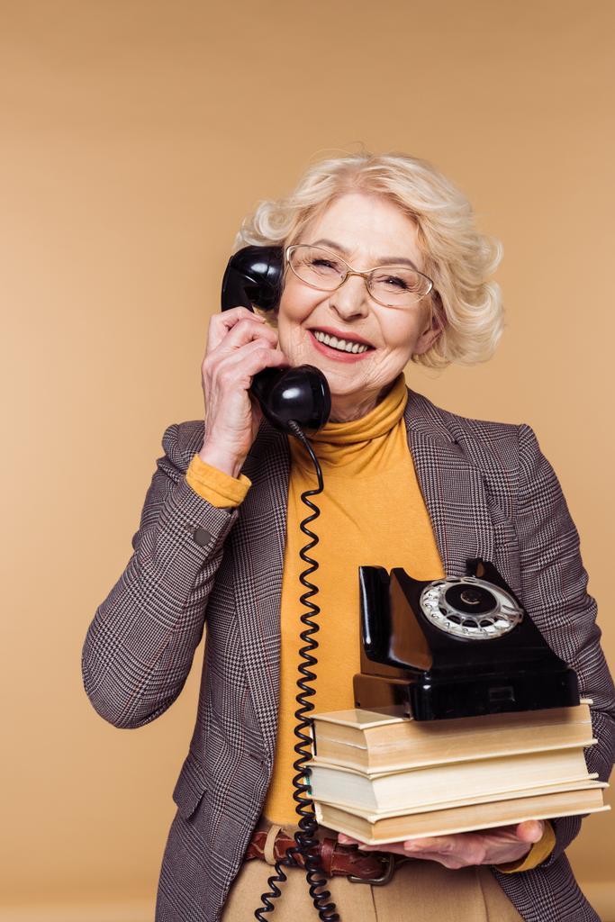 glückliche modische Seniorin mit Brille, die am Drehtelefon spricht und einen Stapel Bücher in der Hand hält  - Foto, Bild