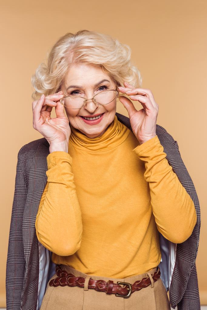 donna anziana sorridente alla moda con giacca sulle spalle che regola gli occhiali isolati su sfondo beige
  - Foto, immagini