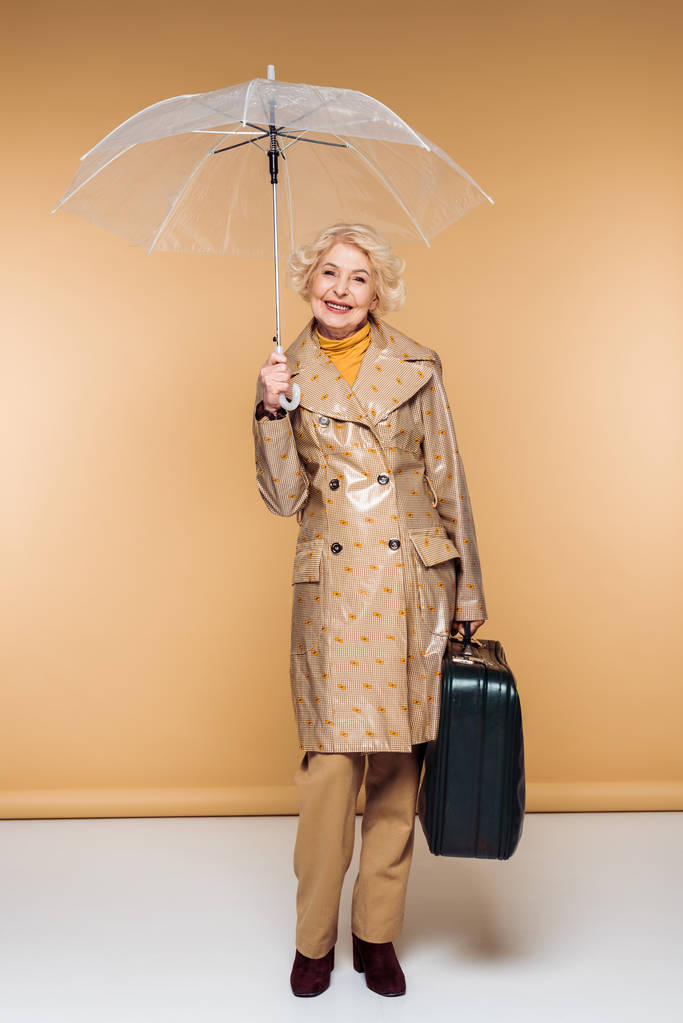 модна старша жінка-мандрівник в траншеї пальто з парасолькою і валізою
  - Фото, зображення