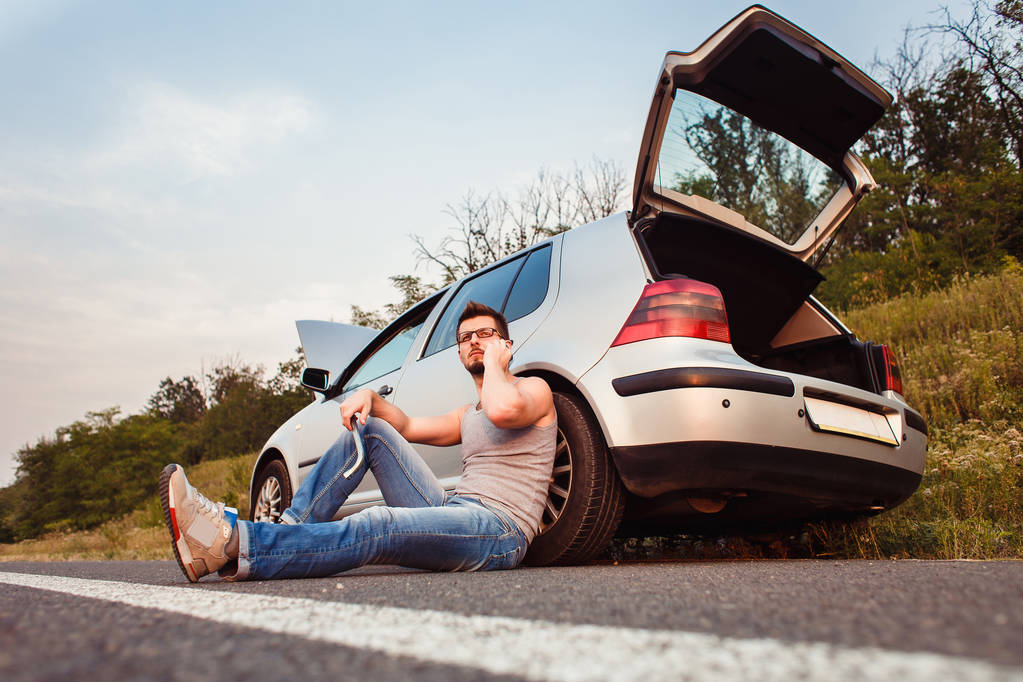 A young man sits on the asphalt, near his broken car and call technical support. Damage on the road - Photo, Image