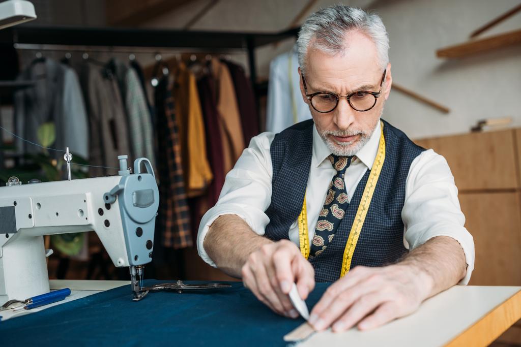 Senior kleermaker overzicht puttend uit doek op naaien workshop - Foto, afbeelding