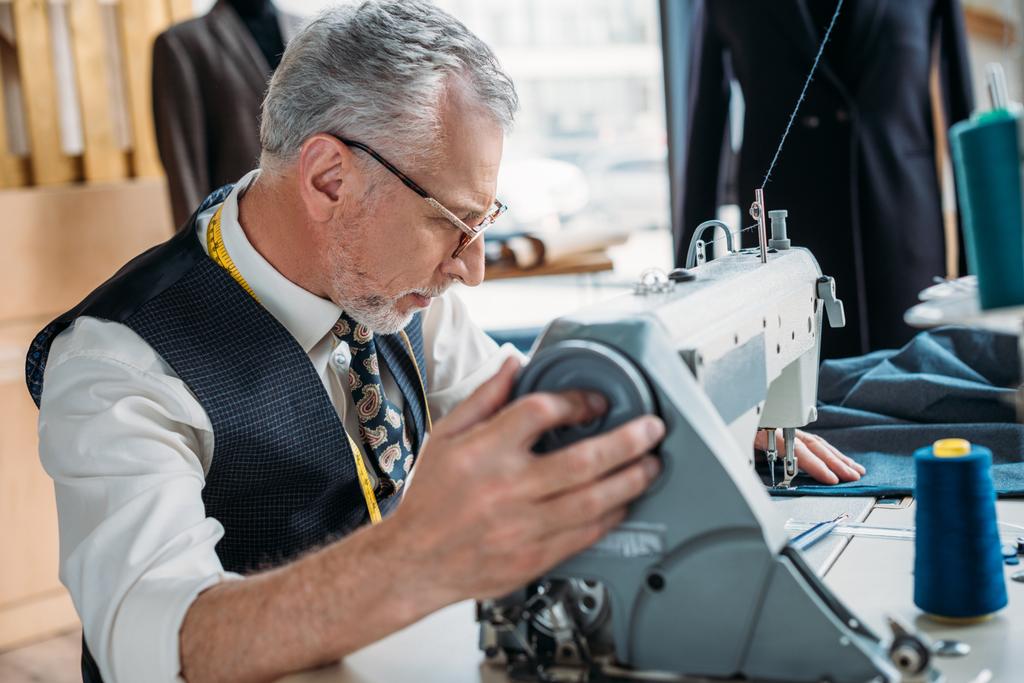 Zijaanzicht van senior kleermaker naaien doek met naaimachine op naaien workshop - Foto, afbeelding